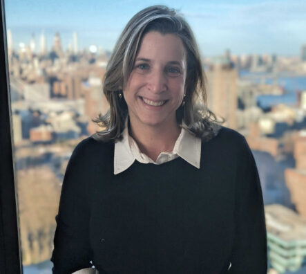 Rebecca Katz stands in front of a window with the New York City skyline in the background.