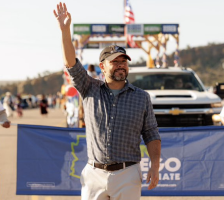 Ruben Gallego waves a hand to a crowd at the Navajo Nation Parade. He is wearing a black Marines cap.