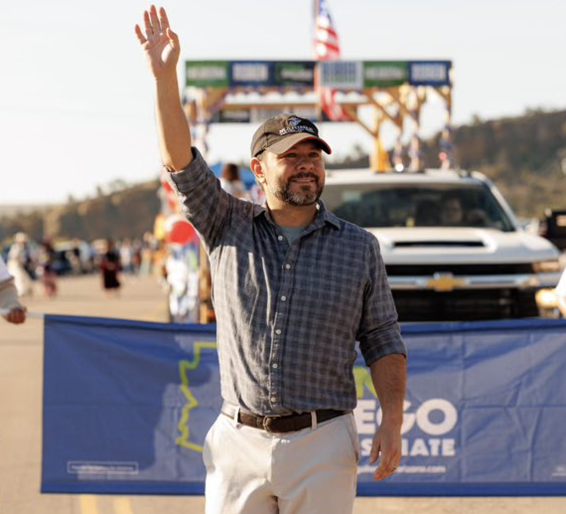 Ruben Gallego waves a hand to a crowd at the Navajo Nation Parade. He is wearing a black Marines cap.